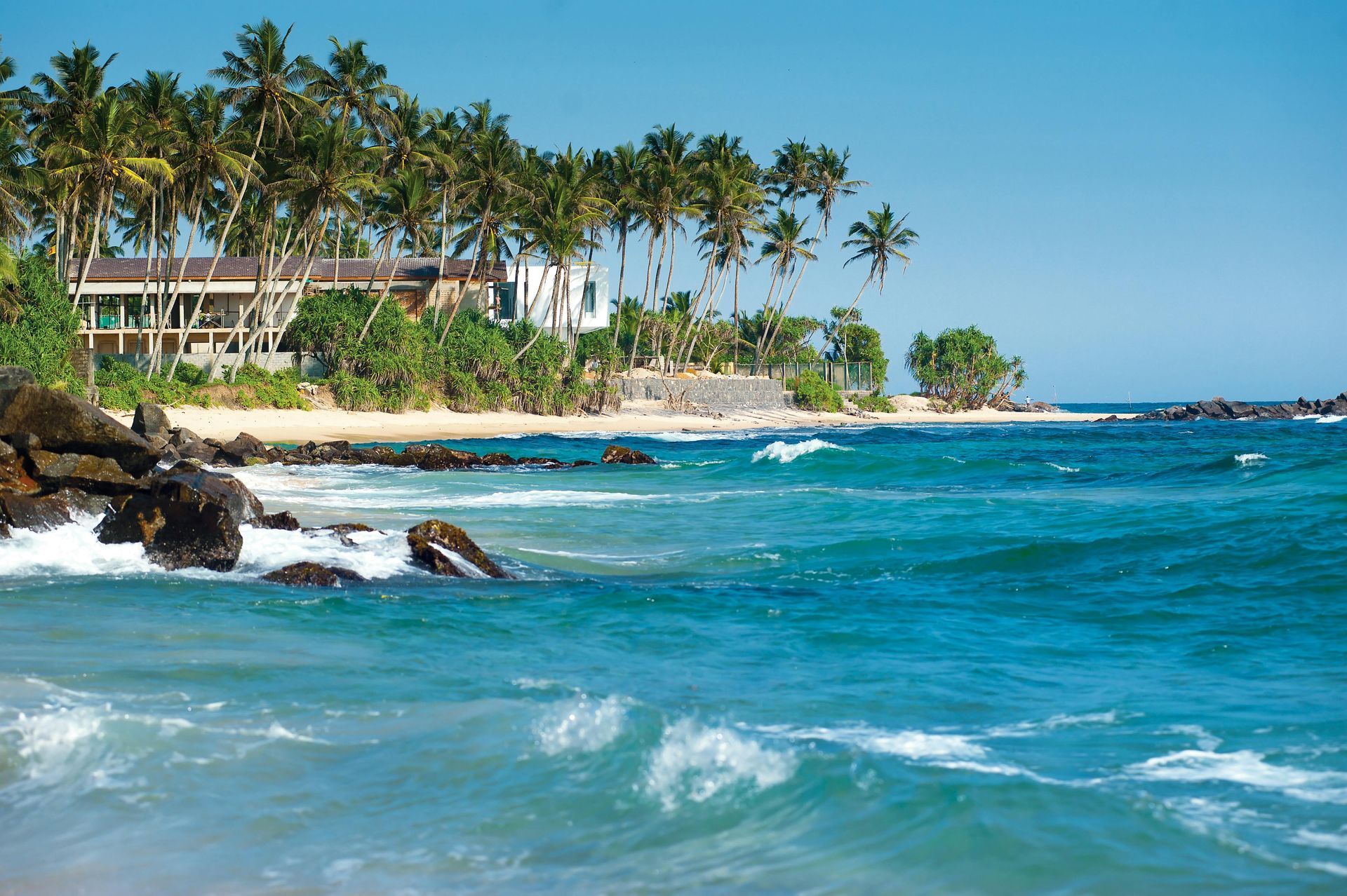 Ocean waves crashing on a rocky shore with palm trees and a building in the background. Bright blue water.