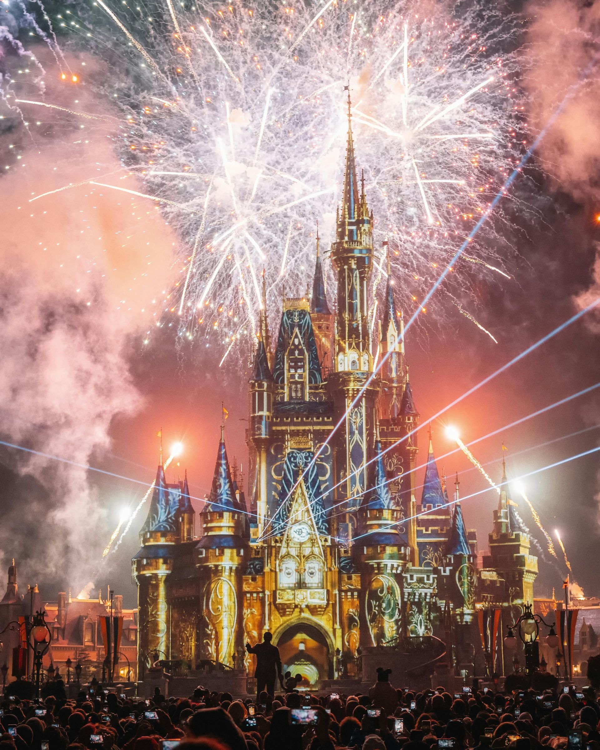 Cinderella Castle lit up with colorful fireworks exploding in the night sky over a large crowd.