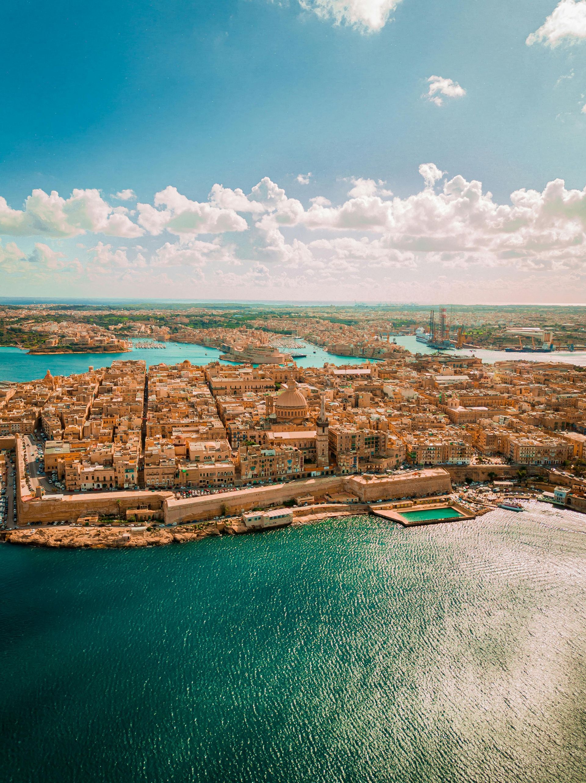 Aerial view of Valletta, Malta, a historic city with tan buildings surrounded by blue water under a partly cloudy sky.