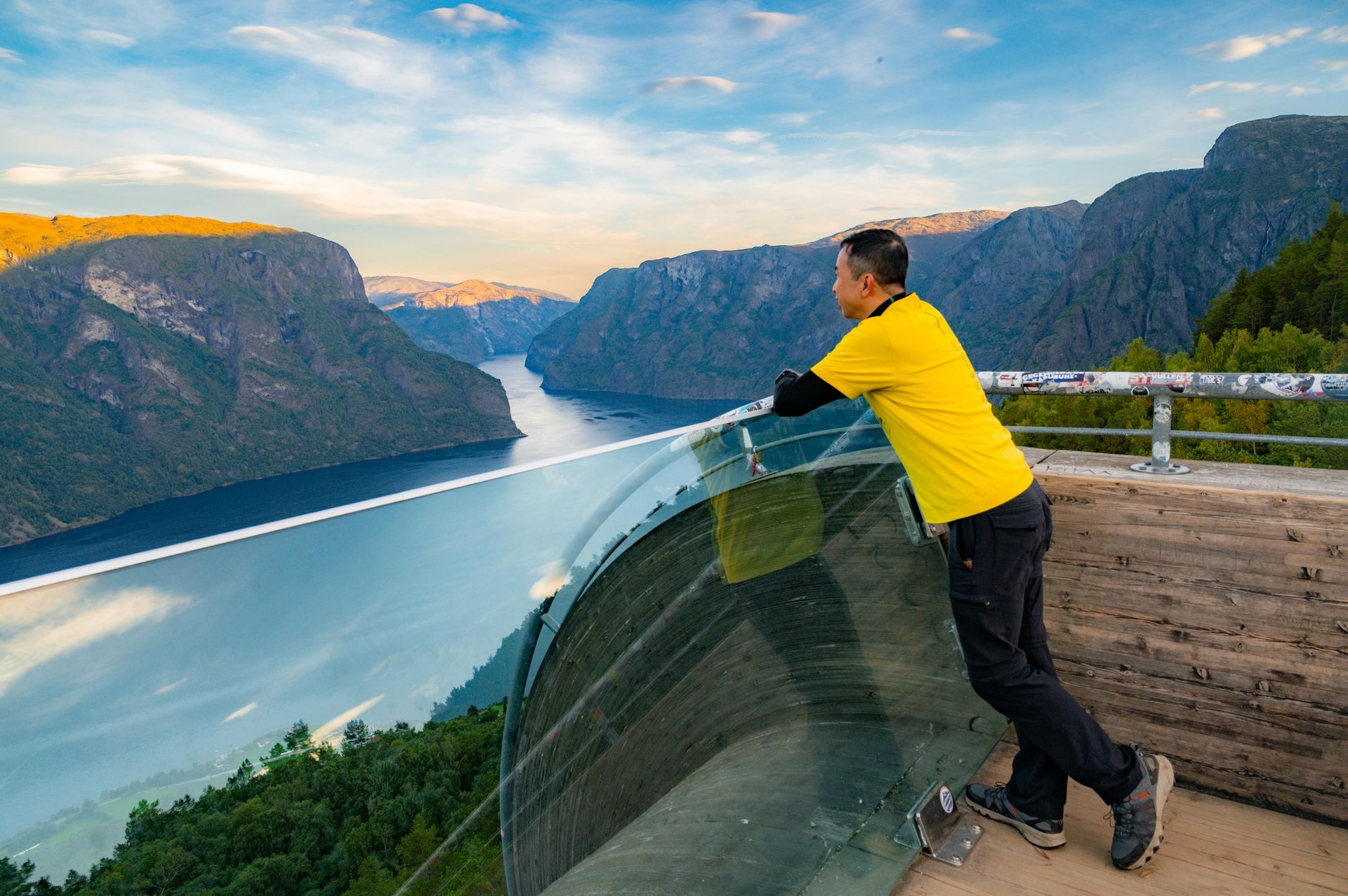 Man in yellow shirt looks at a fjord from a glass viewing platform.