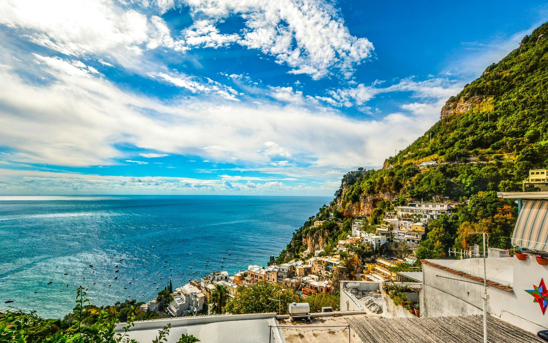 Coastal Italian village nestled on hillside, overlooking the sea under a bright, partly cloudy sky.