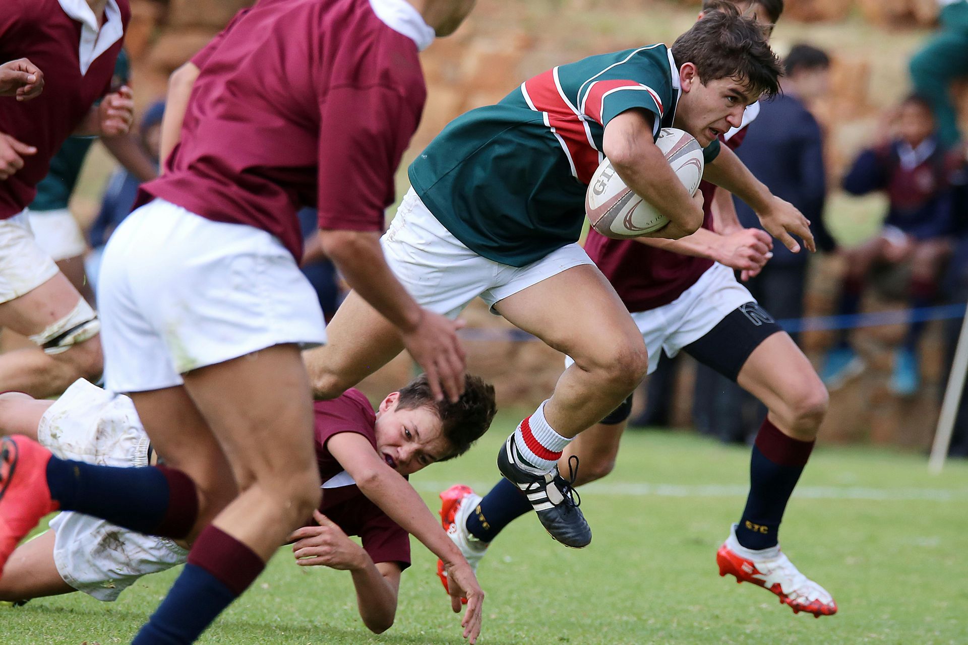 Rugby player in green jersey runs with ball, tackled by players in maroon. Outdoor field.