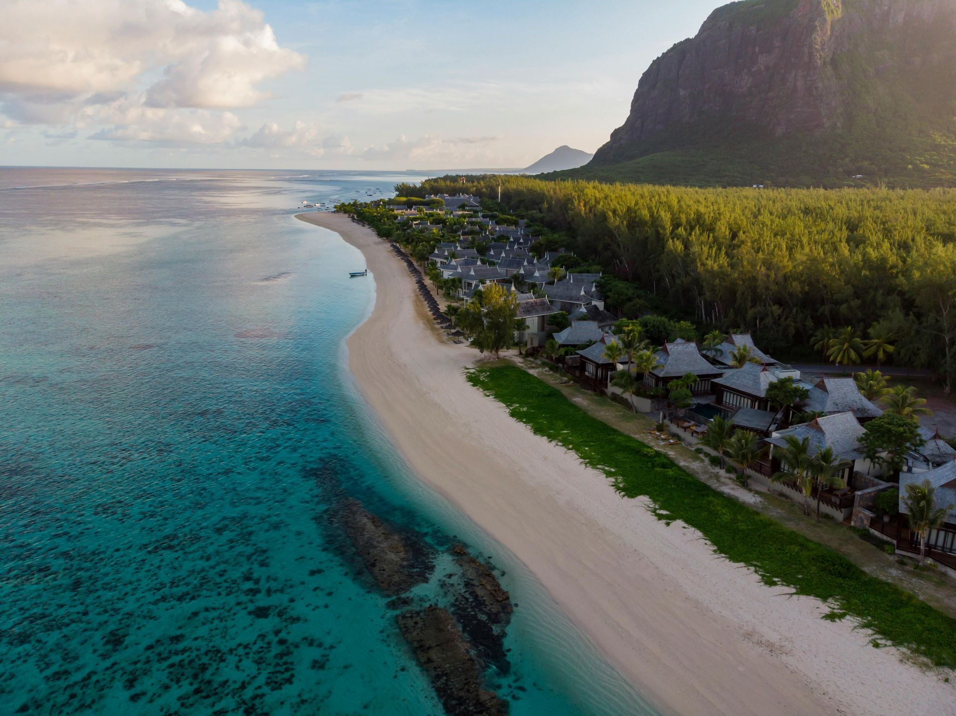 Aerial view of a white sandy beach with turquoise water, a resort, and a green forested area at the base of a mountain.