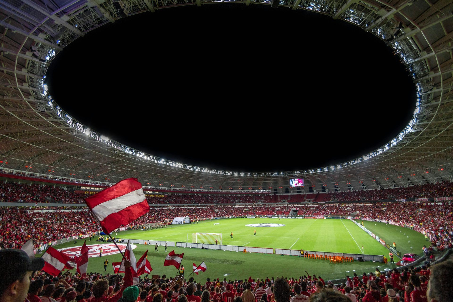Soccer stadium at night, red and white flags waving, cheering crowd, illuminated field, open roof.