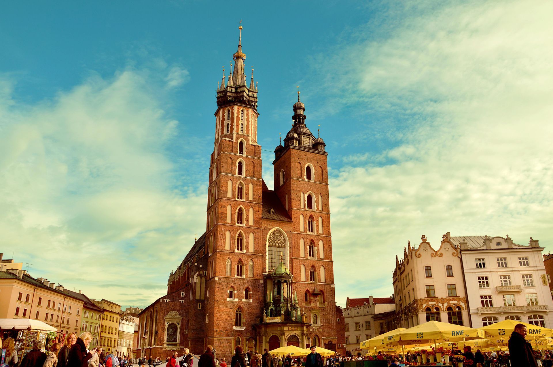 St. Mary's Basilica in Kraków, Poland, with tall brick towers, blue sky, and yellow umbrellas in the square.