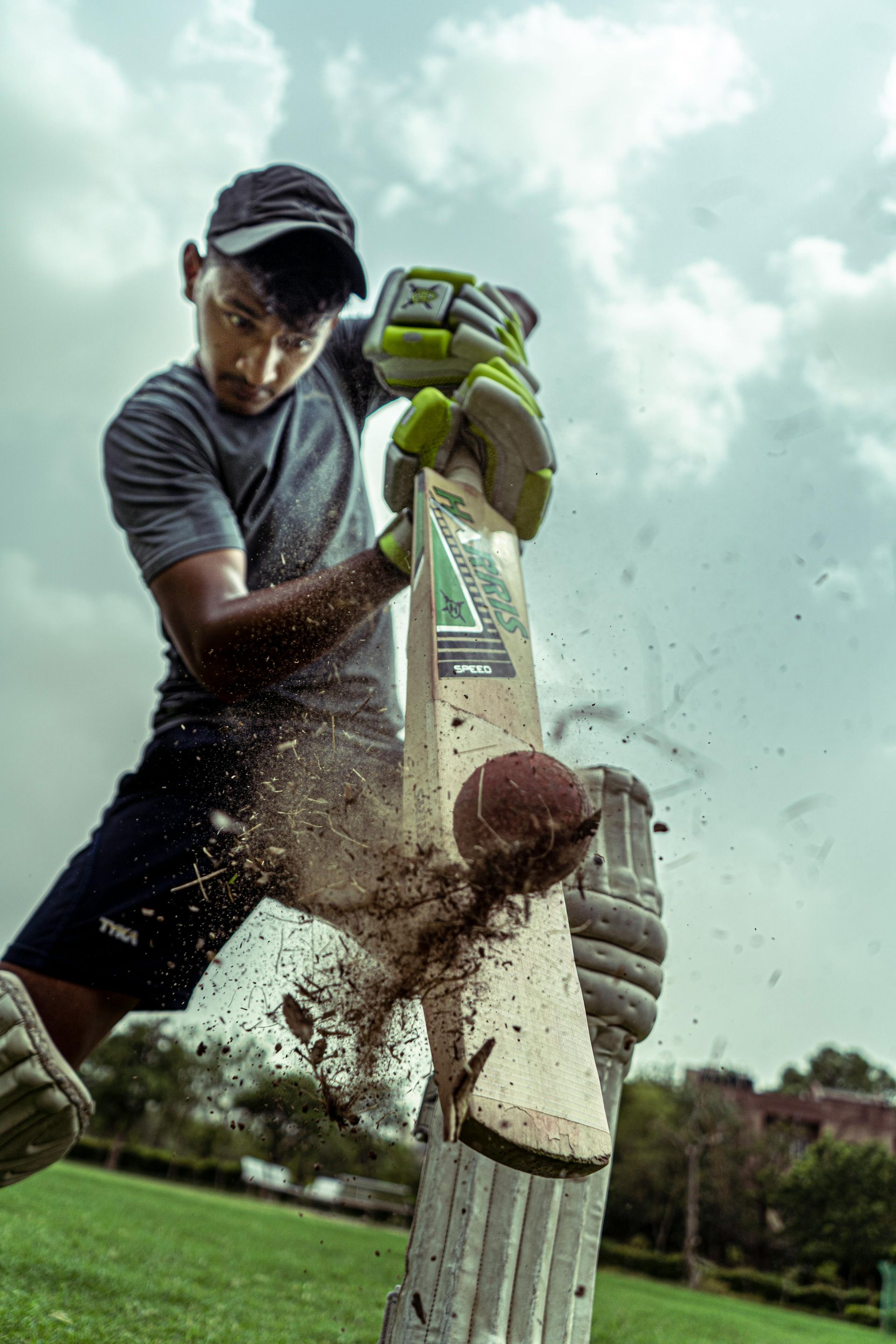 Cricketer swings bat, hitting the ball, sending dirt flying. Cloudy sky, green grass.