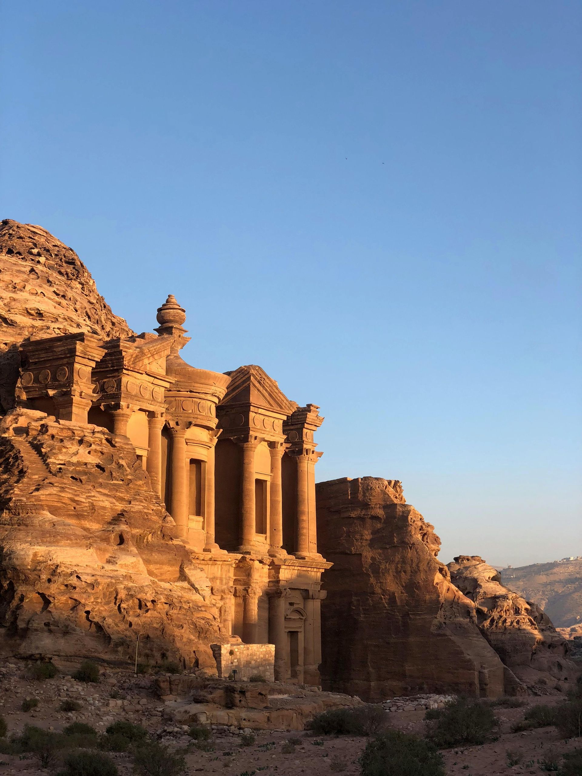 The Monastery in Petra, Jordan, carved into sandstone cliffs, bathed in warm sunlight against a clear blue sky.