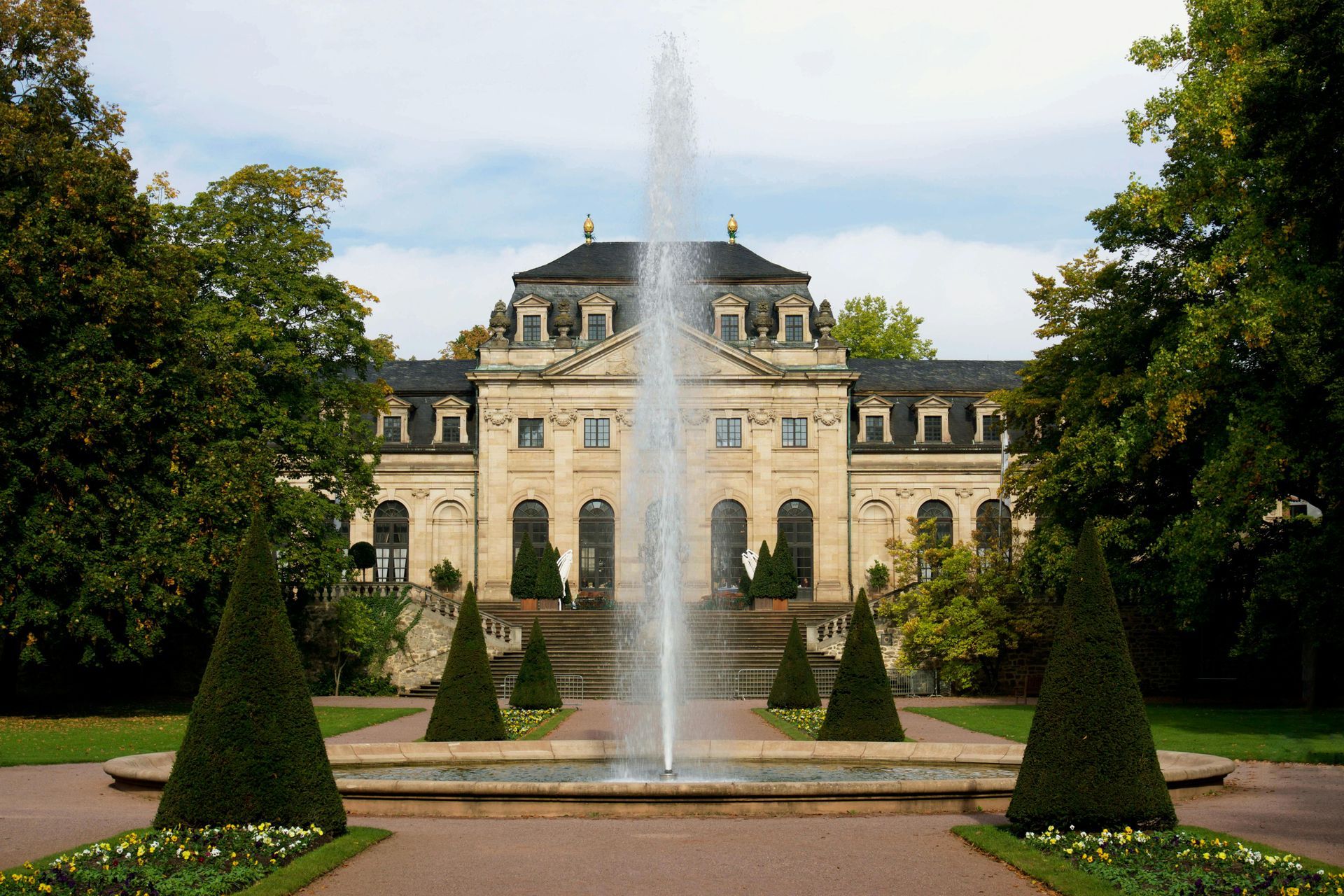 Baroque palace with fountain in front, surrounded by trees and a garden path.