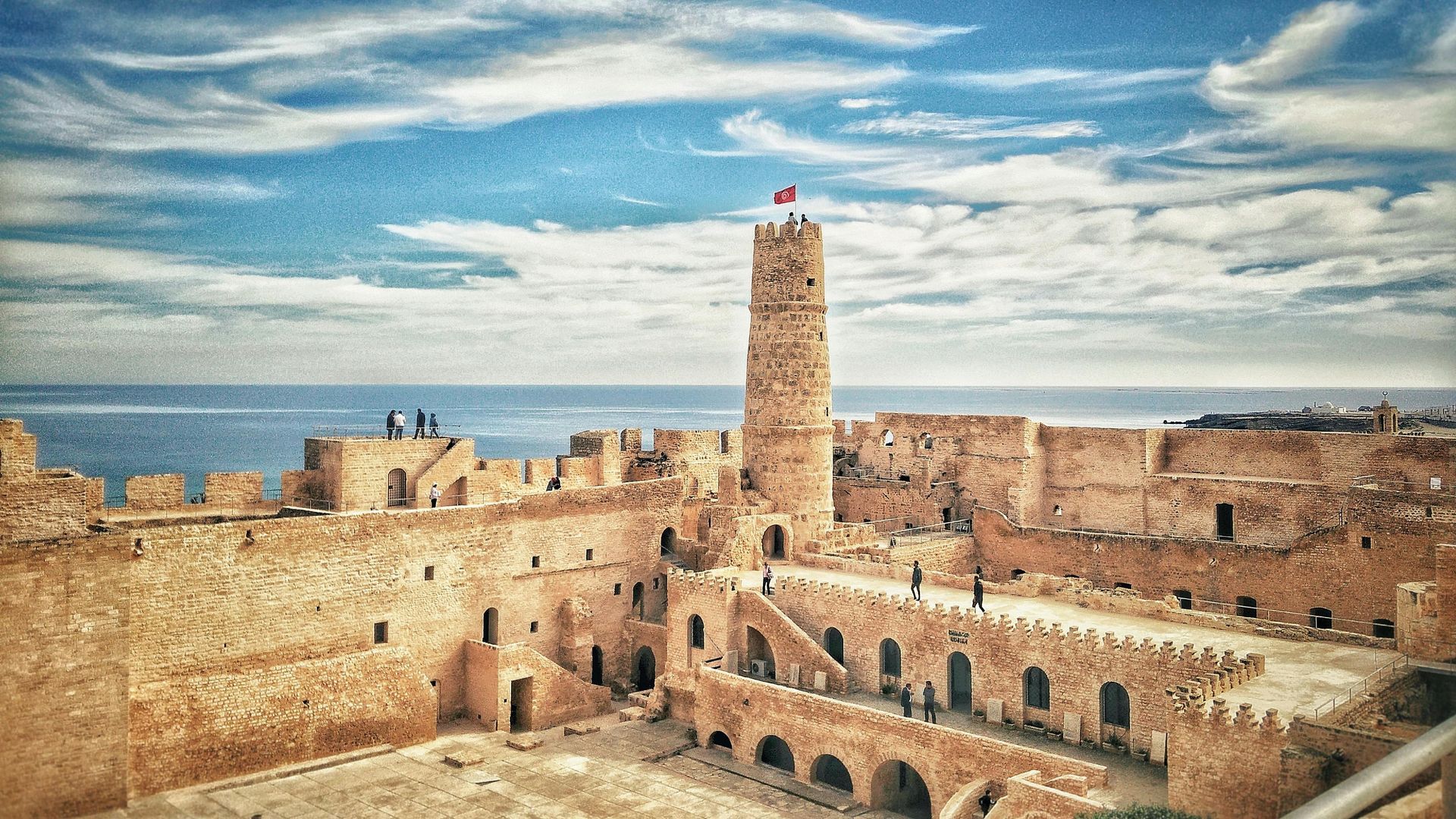 Fortified stone structure with a tall tower, overlooking the ocean under a blue and cloudy sky.
