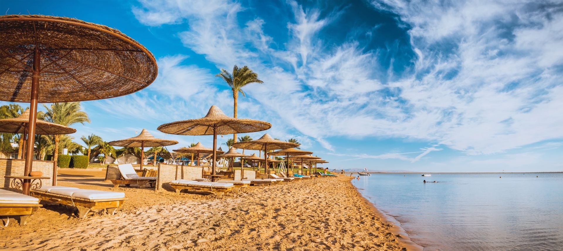 Beach with straw umbrellas, lounge chairs, and calm water under a blue sky with clouds.