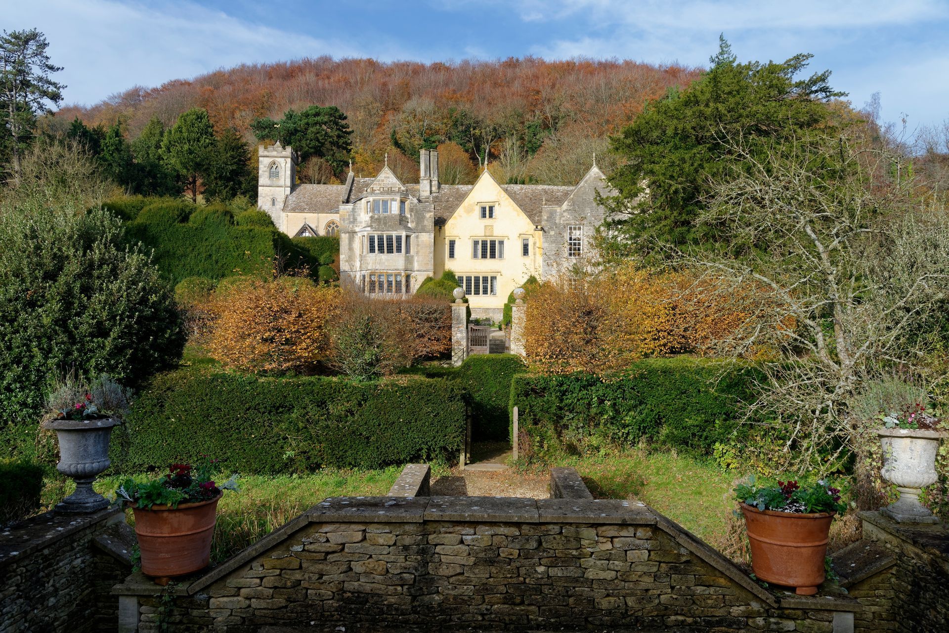 Manor house nestled in autumn-colored trees, viewed from a terraced garden with flower pots.