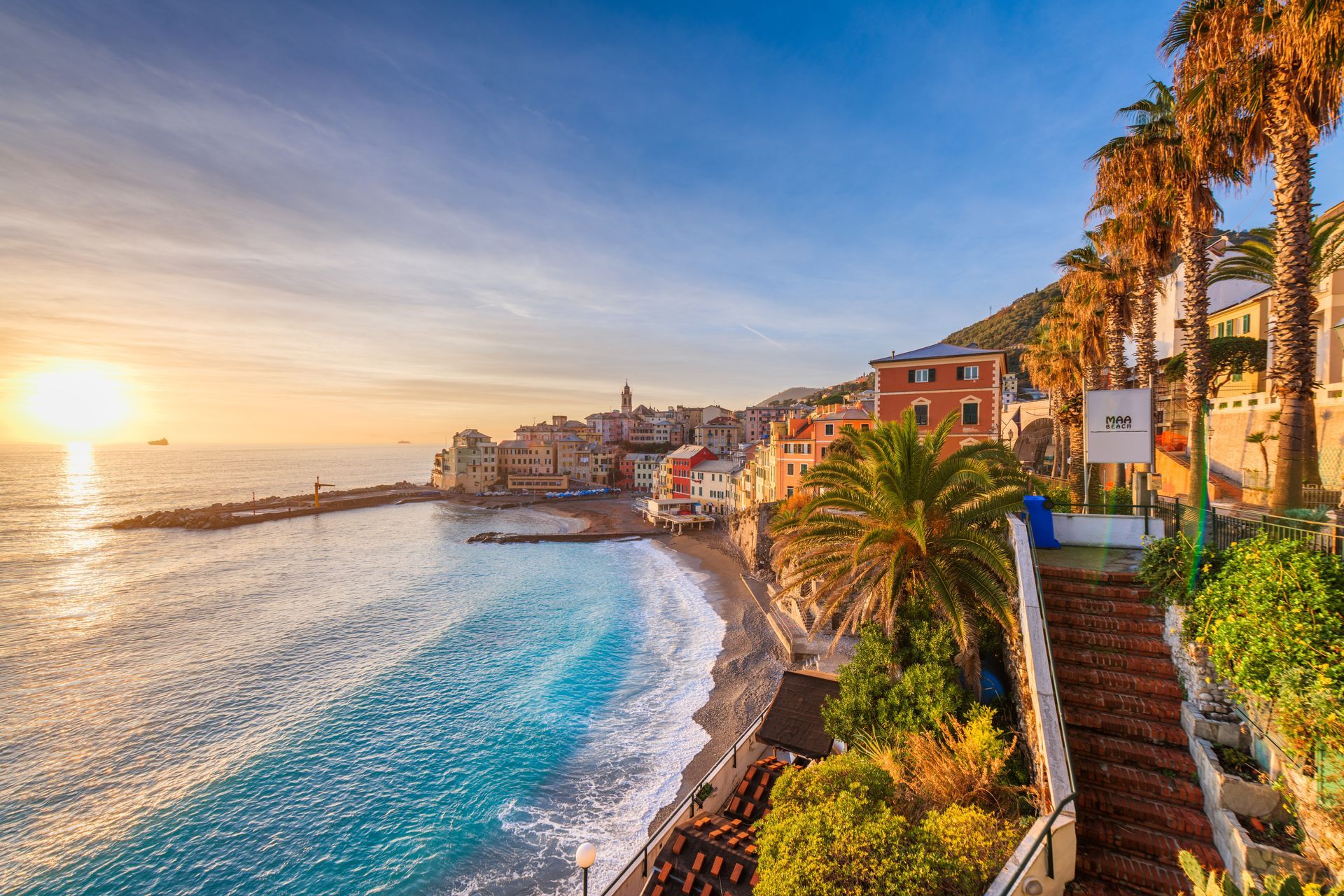 Beach scene at sunset. Turquoise water, buildings on the coast, palm trees, stone steps leading down.