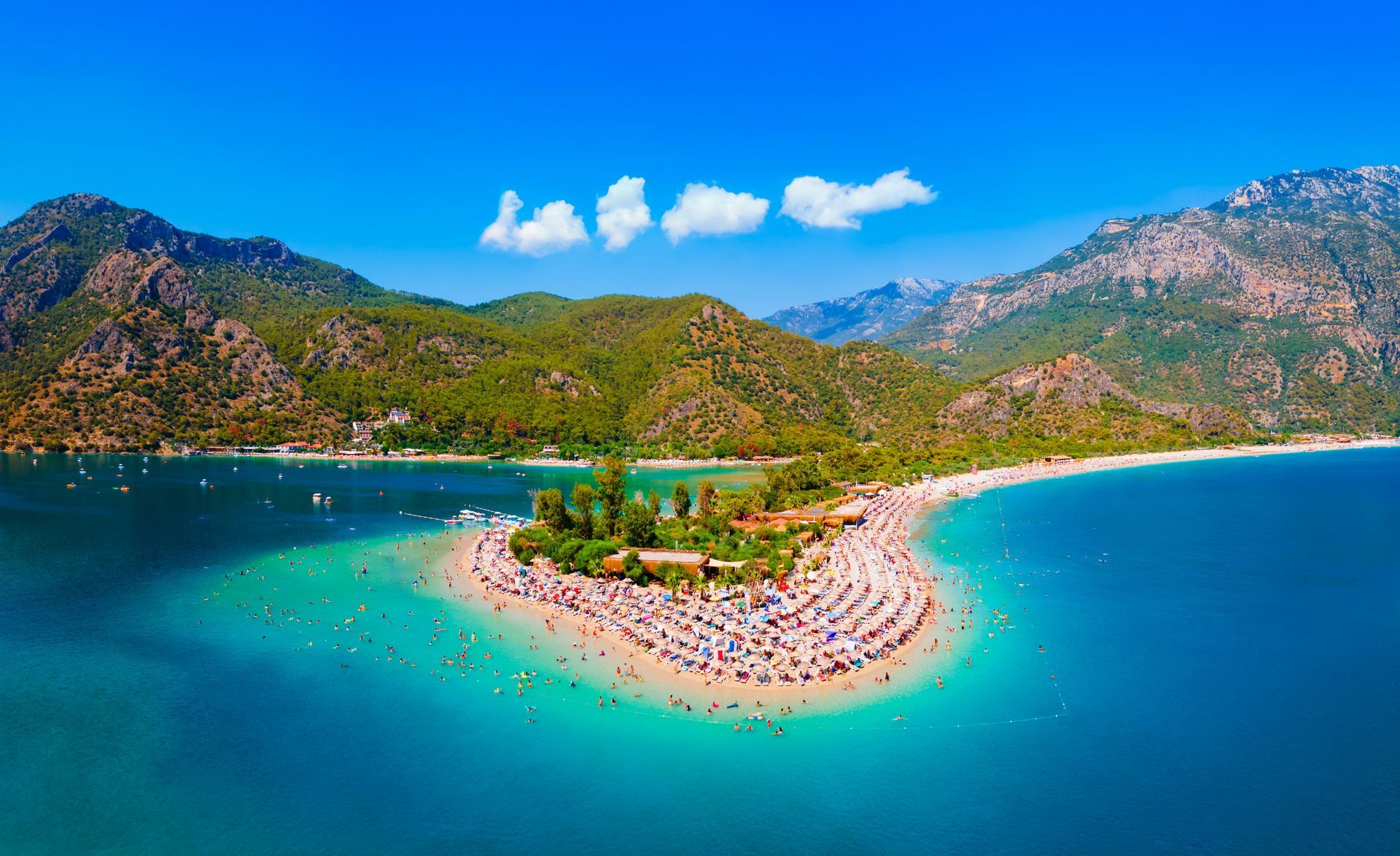 Turquoise water surrounds a sandy beach with many people, mountains in the background under a blue sky.