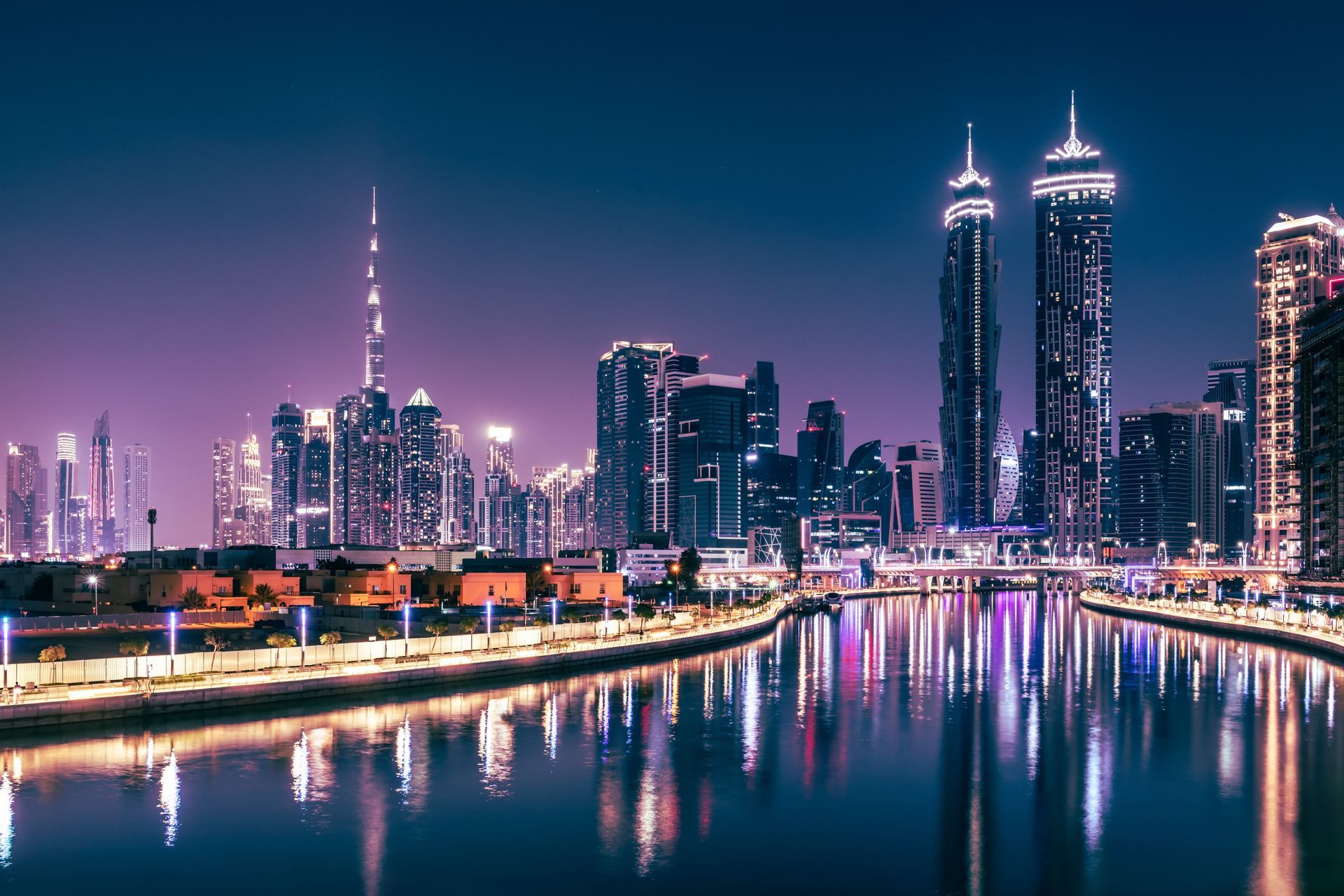 Nighttime cityscape with illuminated skyscrapers reflecting in calm water.