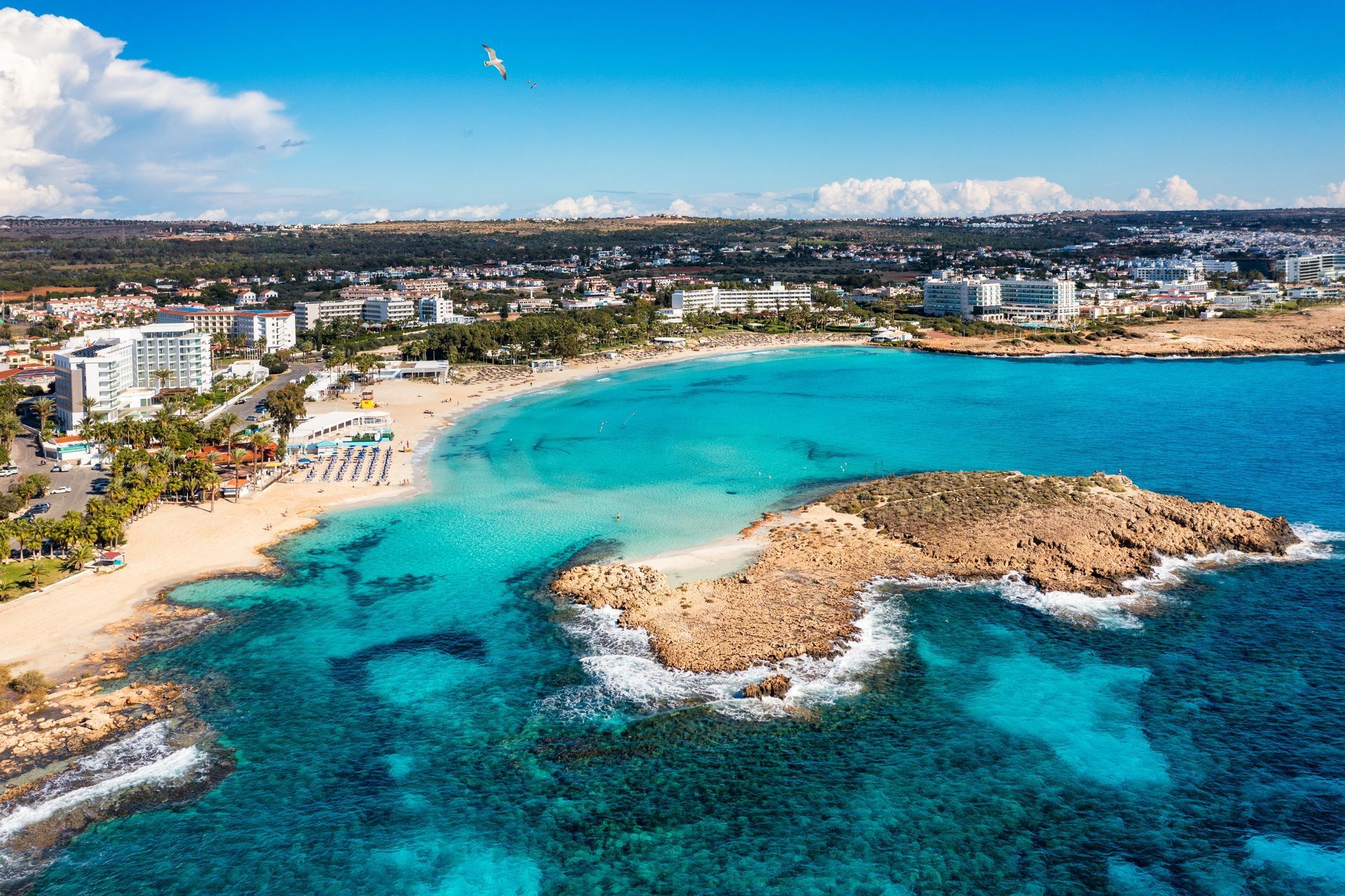 Aerial view of a turquoise bay with sandy beach, rocky island, and coastal buildings under a bright blue sky.