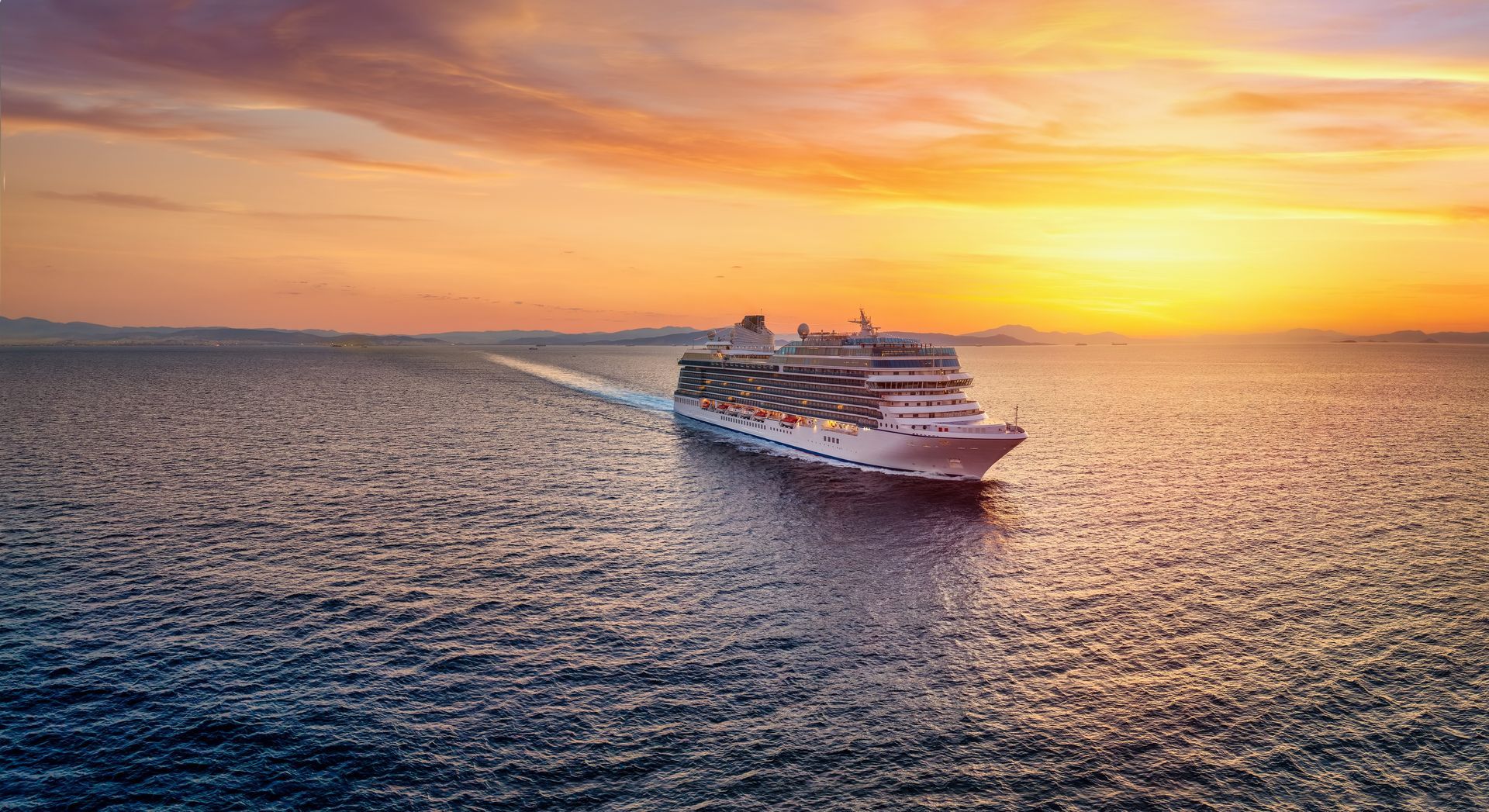 Cruise ship sailing on a calm ocean at sunset; orange and yellow sky, white ship.