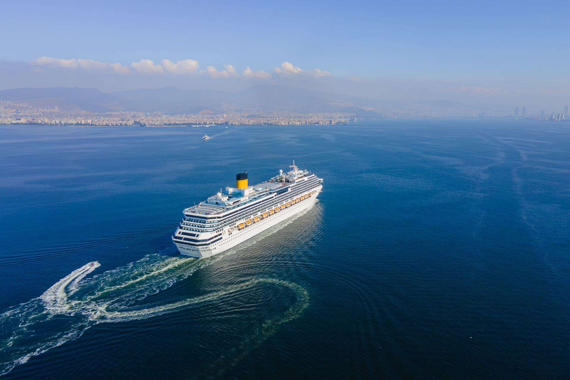Cruise ship sailing on blue water, city skyline in the background.