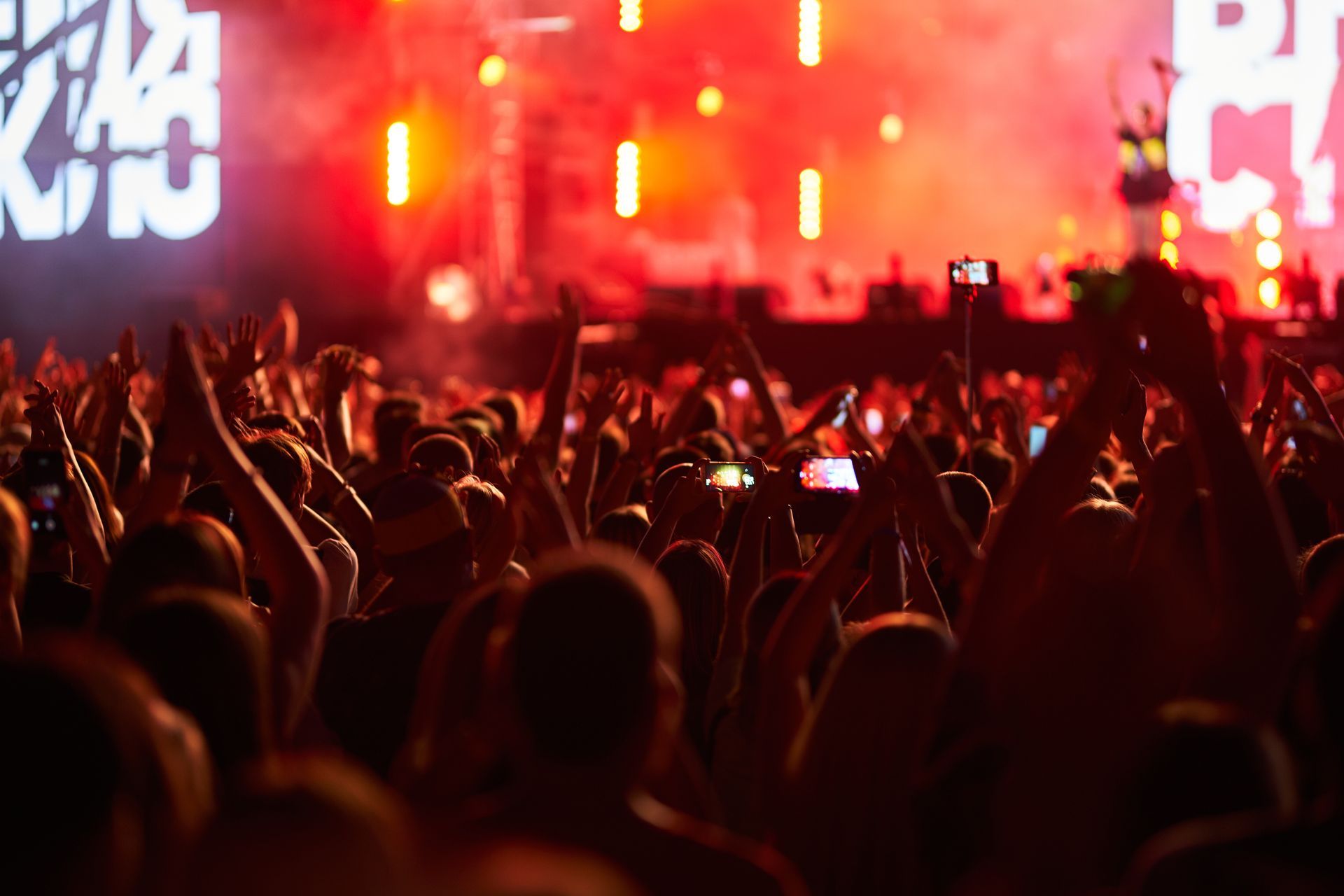 Crowd at a concert with arms raised, illuminated by red and orange stage lights.