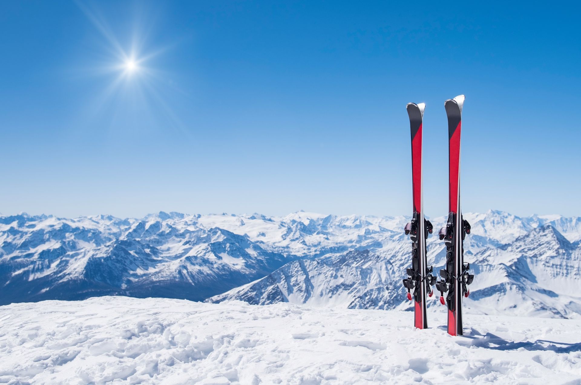 Pair of red skis on snowy mountaintop with sunny blue sky and snow-covered mountains in the distance.