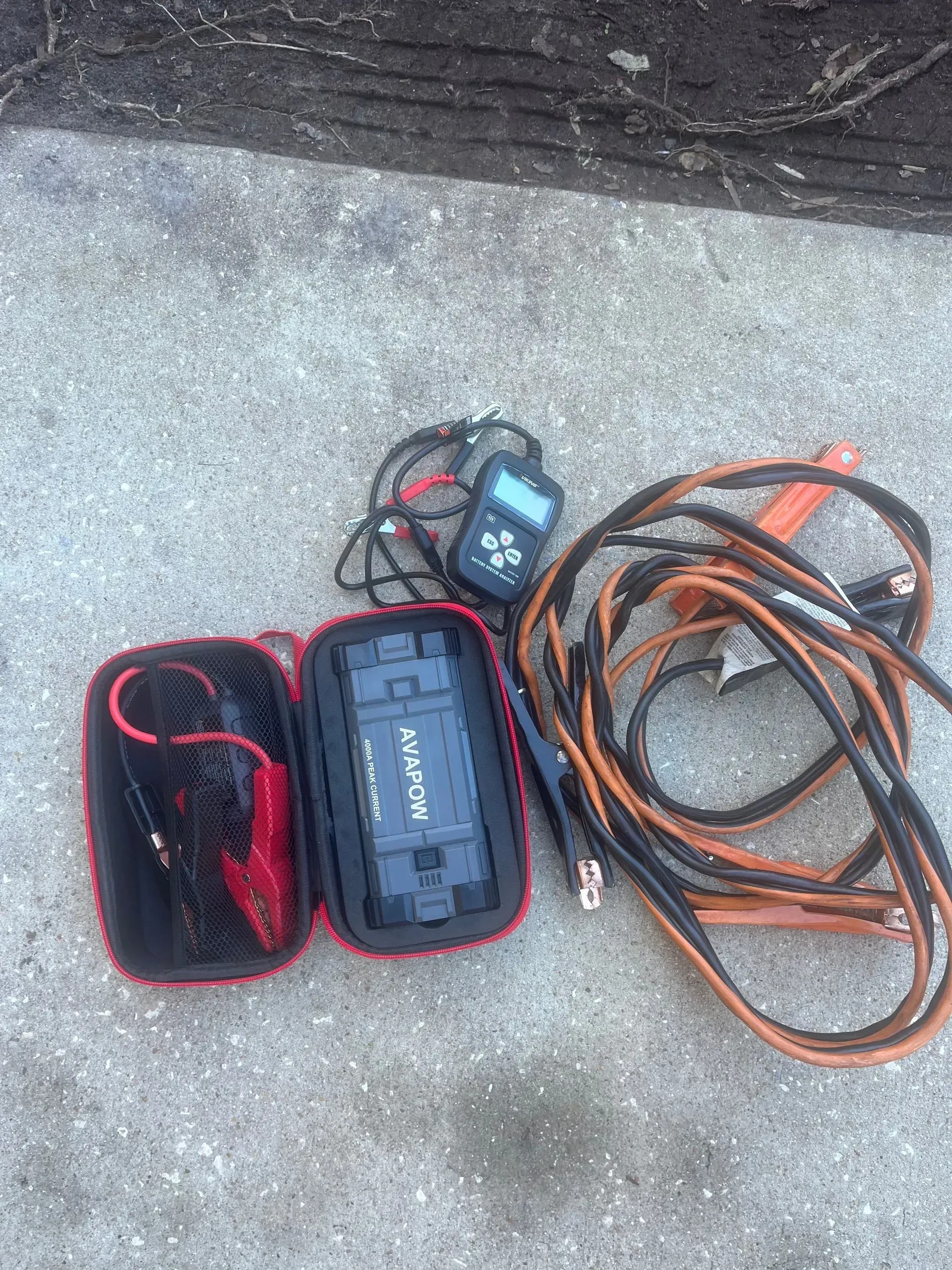 Jump starter kit: black device in a red case, cables, and a battery tester on concrete.