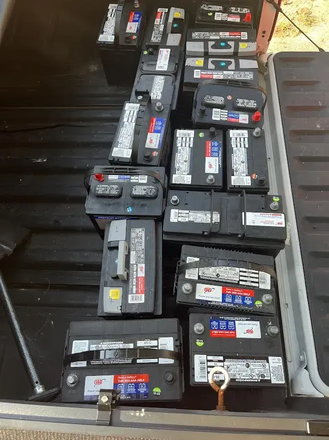 Pile of used car batteries in a truck bed, various brands and sizes, black and gray.