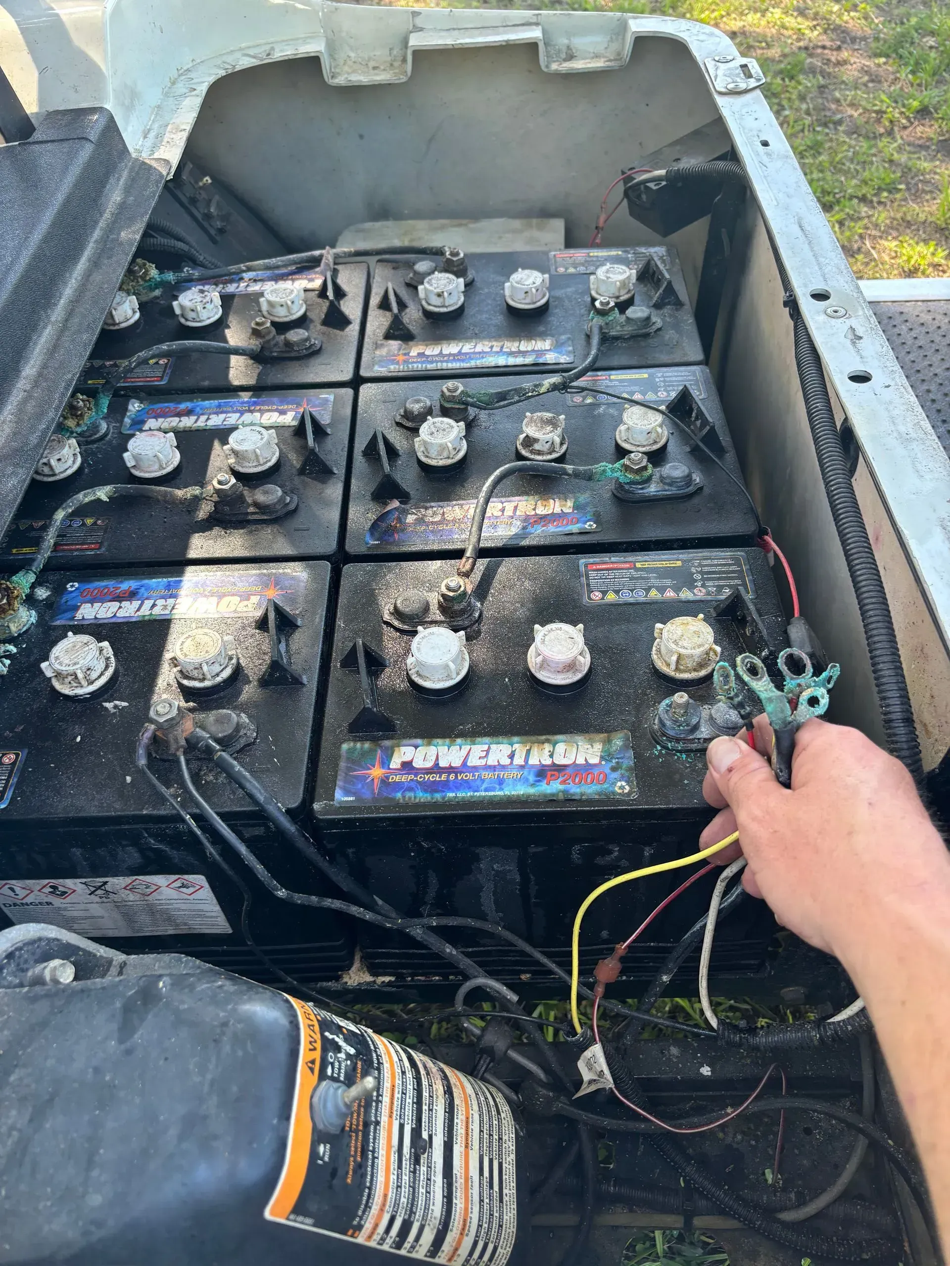 Six golf cart batteries, arranged in a row. A person holds wires. The cart is outdoors.