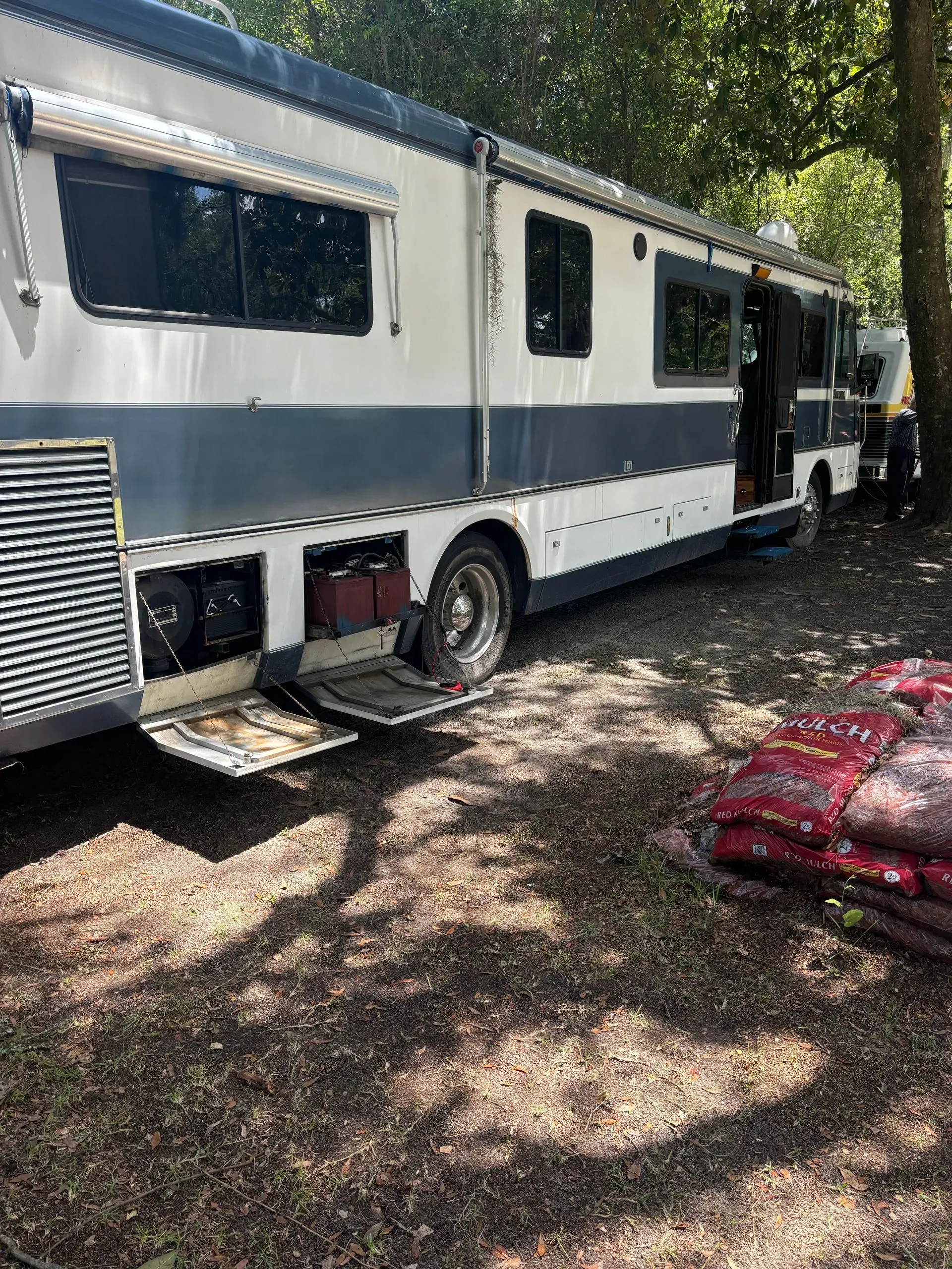 RV parked outdoors, ramp extended. White and blue paint, open door, bags of material nearby.