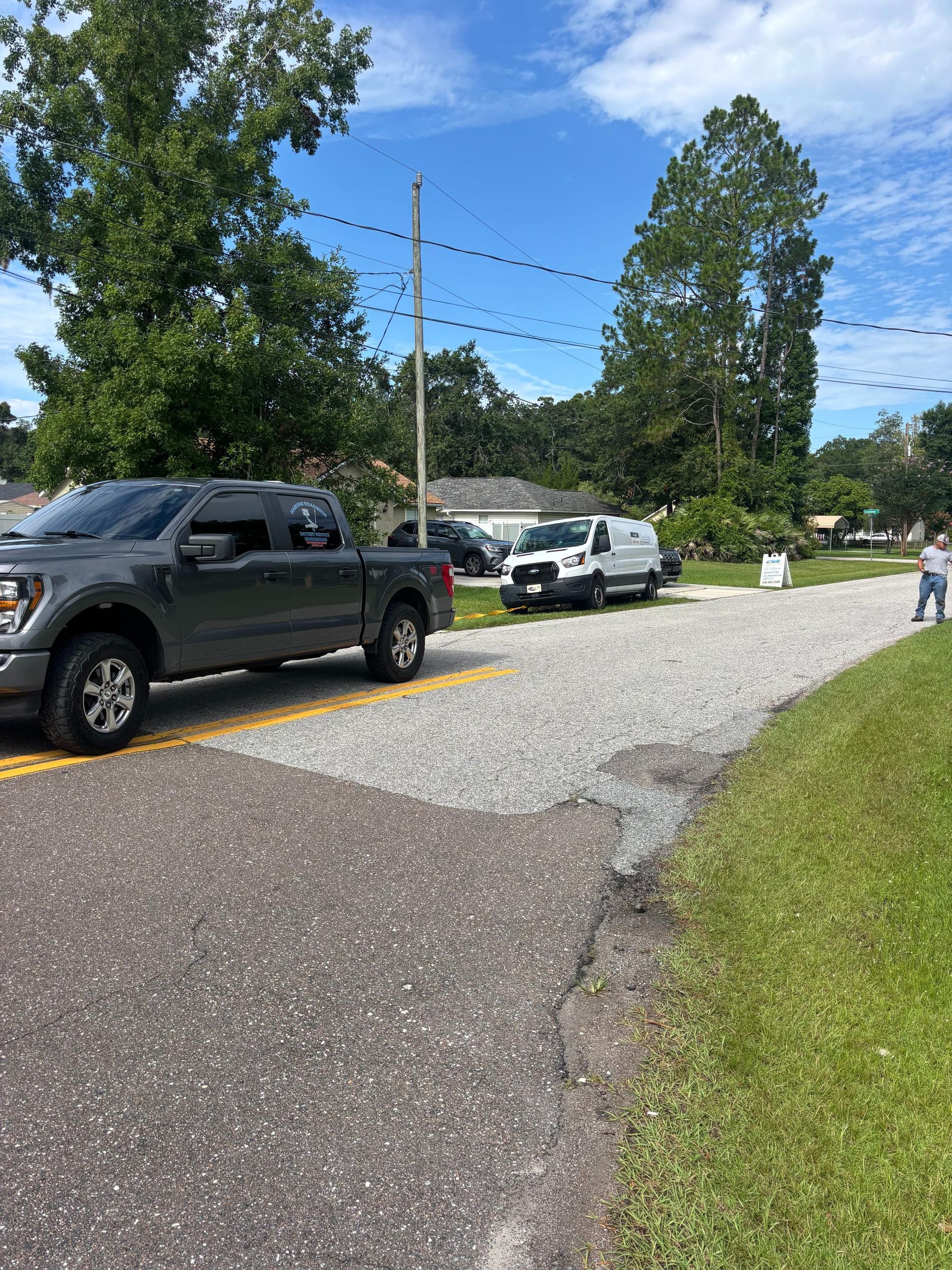 Gray pickup truck on asphalt road, gravel driveway with white van and trees under a blue sky.