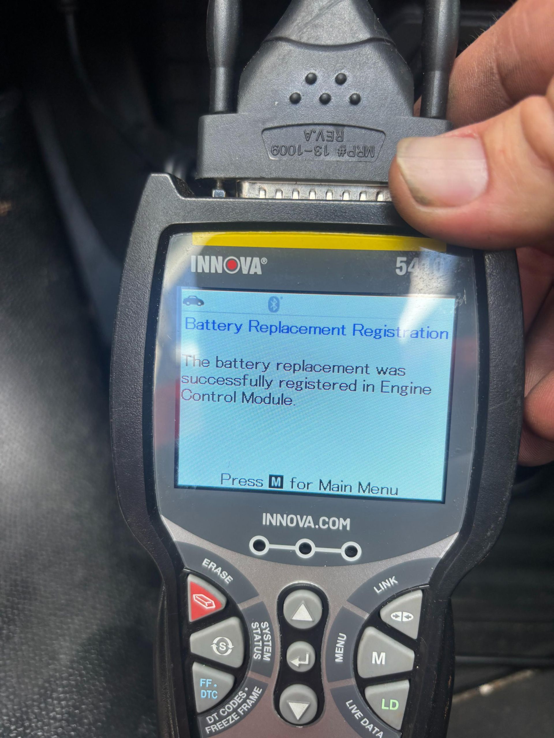 Hand holding a black and gray handheld car diagnostic tool, screen displaying a battery replacement notification.