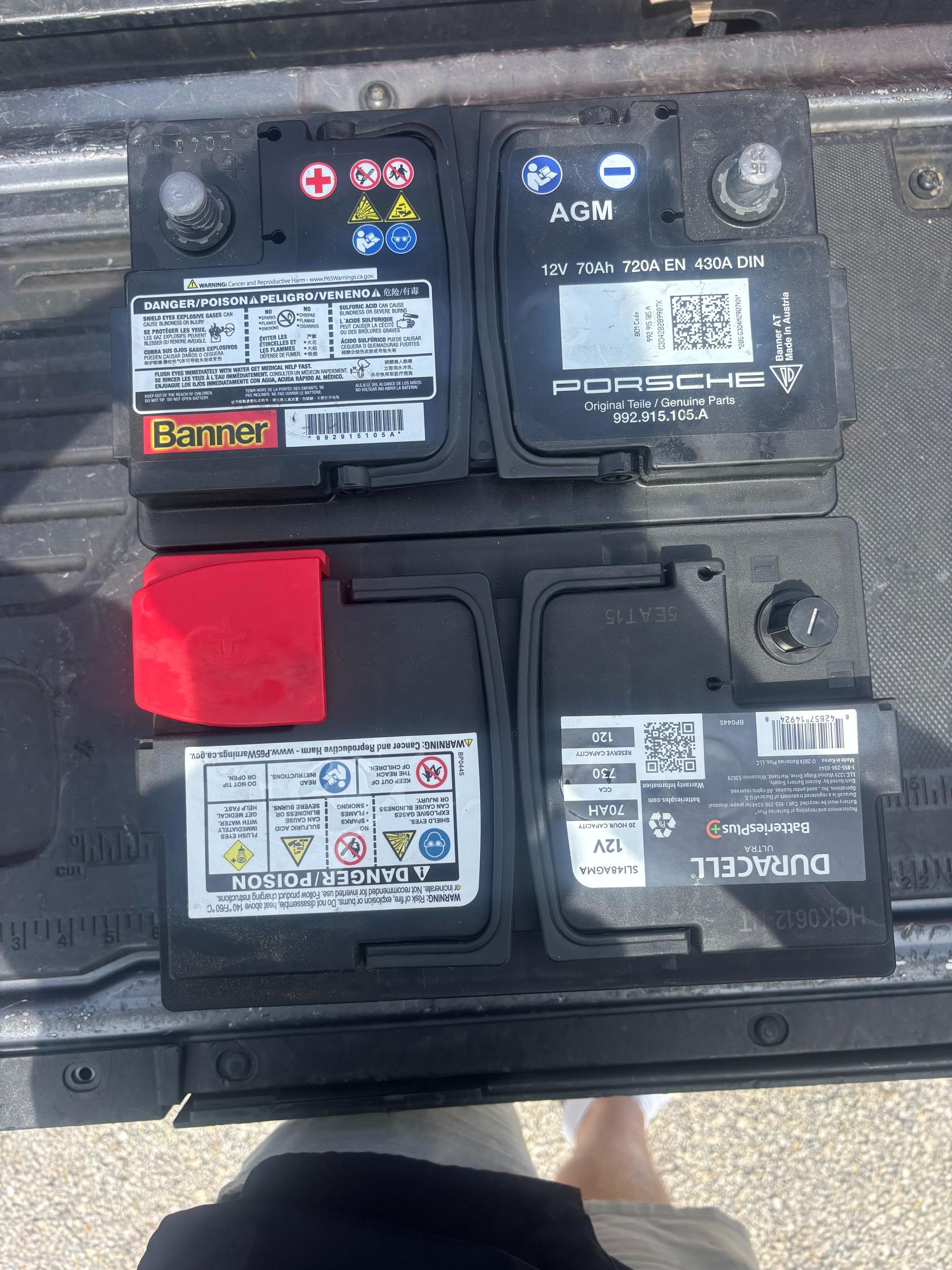 Four car batteries: two black with labels, one with a red cover, sitting on a dark surface.