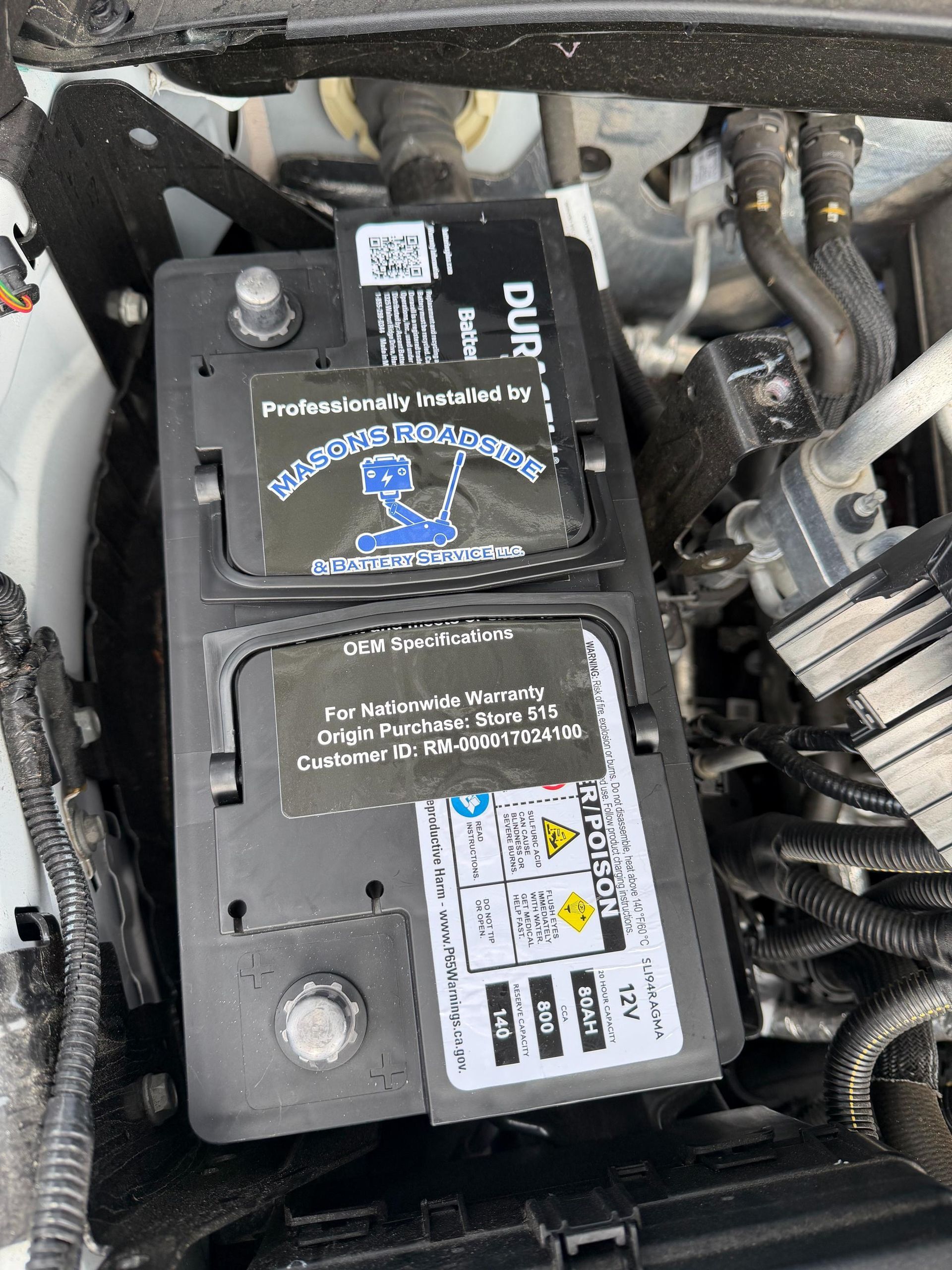 Car battery in engine compartment with warning stickers and black plastic cover.