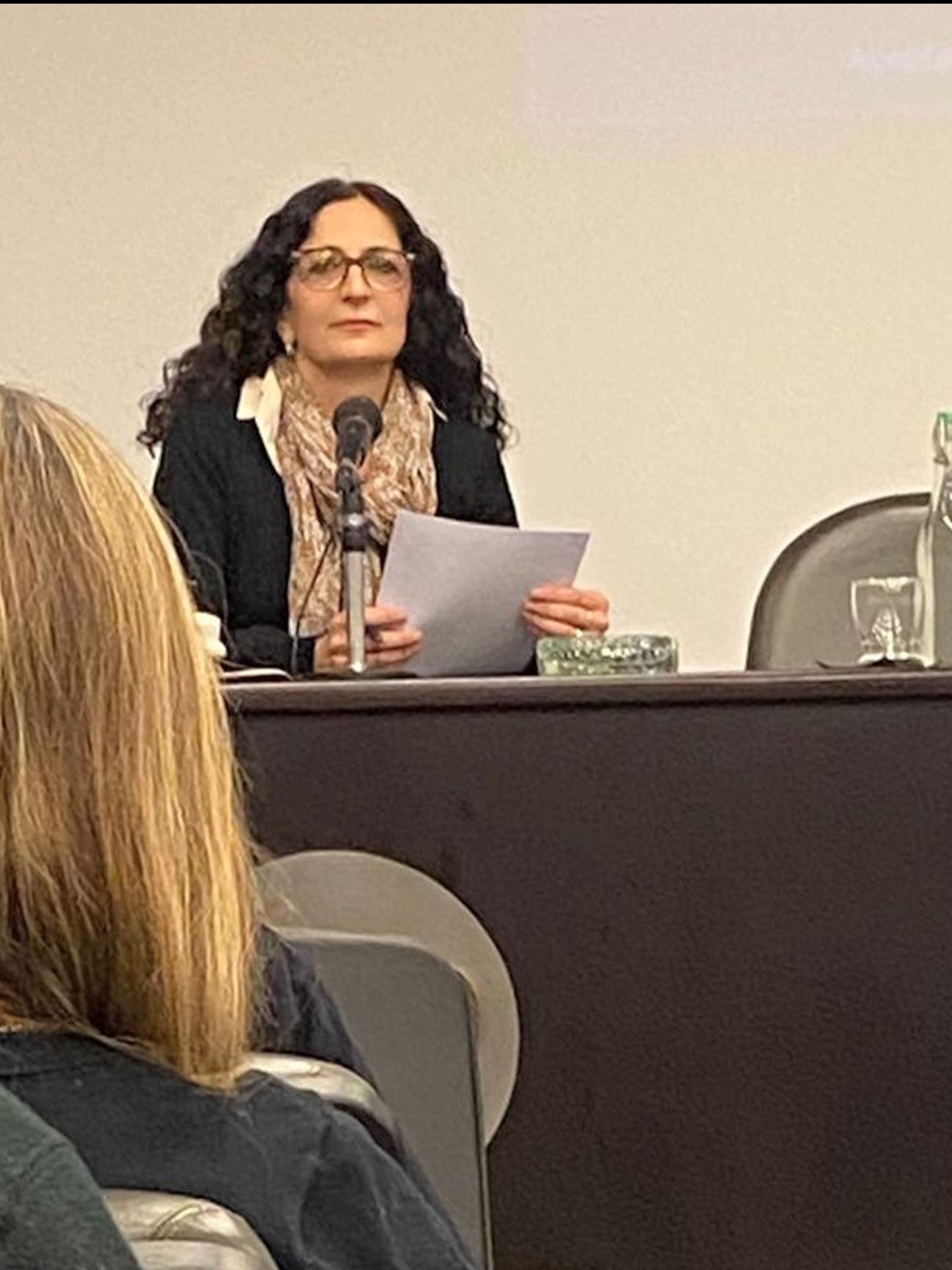 Una mujer está sentada en una mesa sosteniendo un trozo de papel frente a un micrófono.