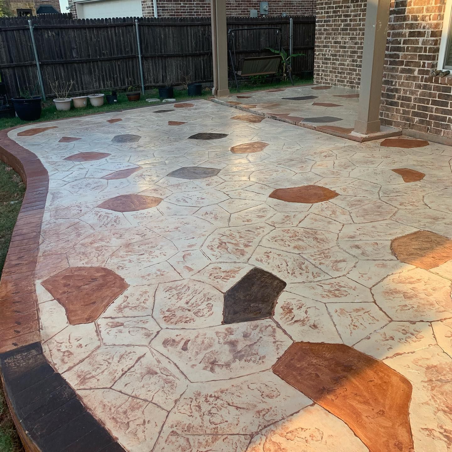 Stamped concrete patio with brown and beige stone pattern, adjacent to a brick building and wooden fence.