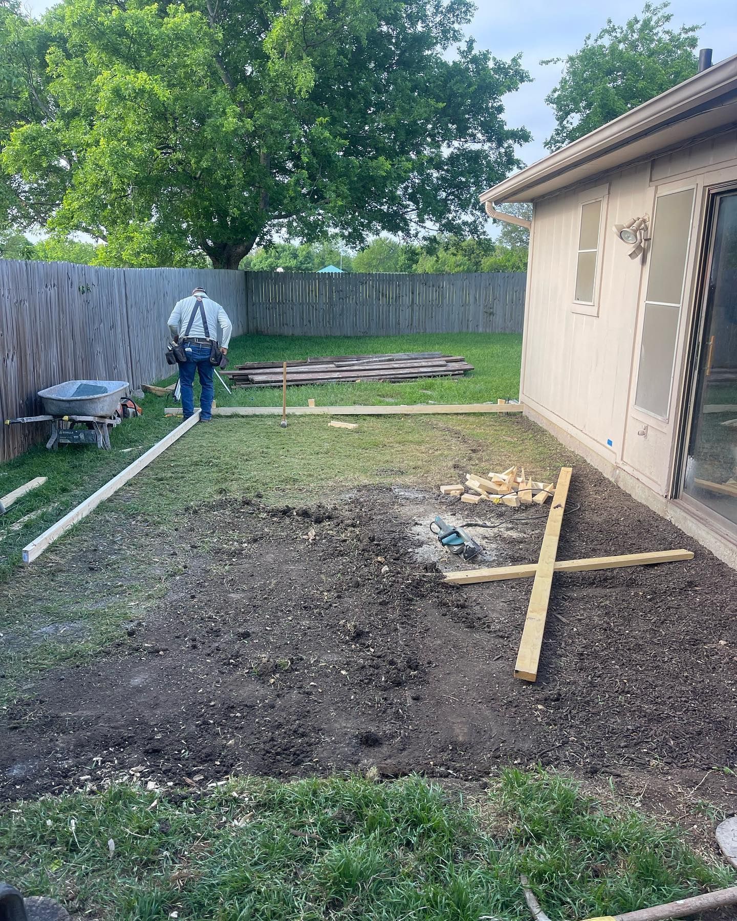 Person building a wooden deck in a backyard with a wooden fence and a house. Dirt and grass visible.