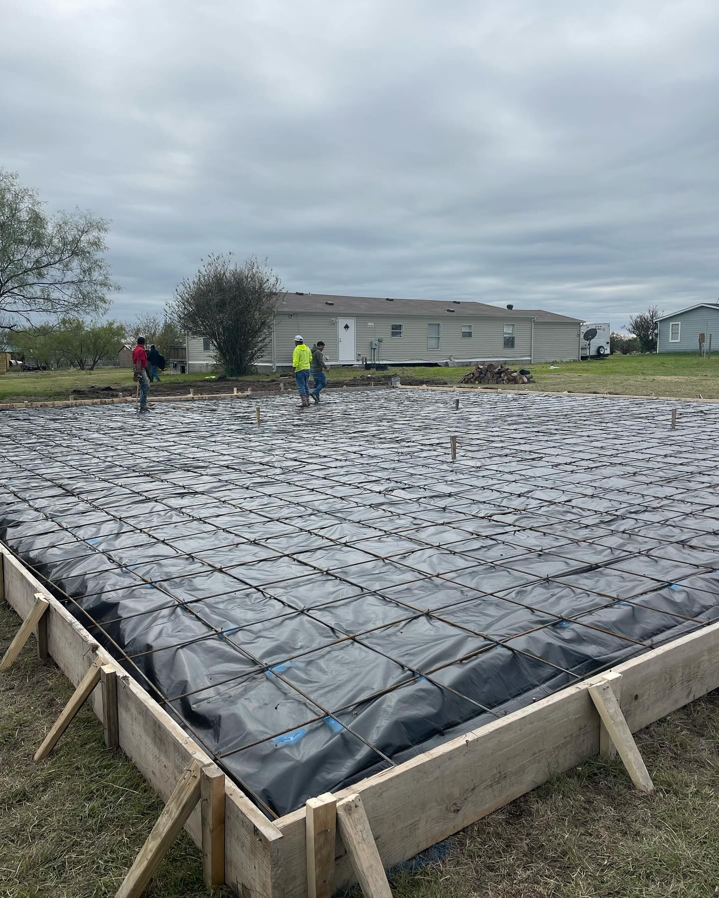 Workers preparing a concrete slab for pouring, wooden frame, black plastic sheet, rebar grid, cloudy day.