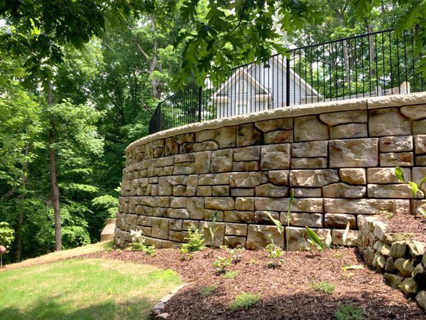 A large stone wall is surrounded by trees and a house in the background.