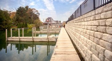 A large brick wall along a lake with a dock and a house in the background.