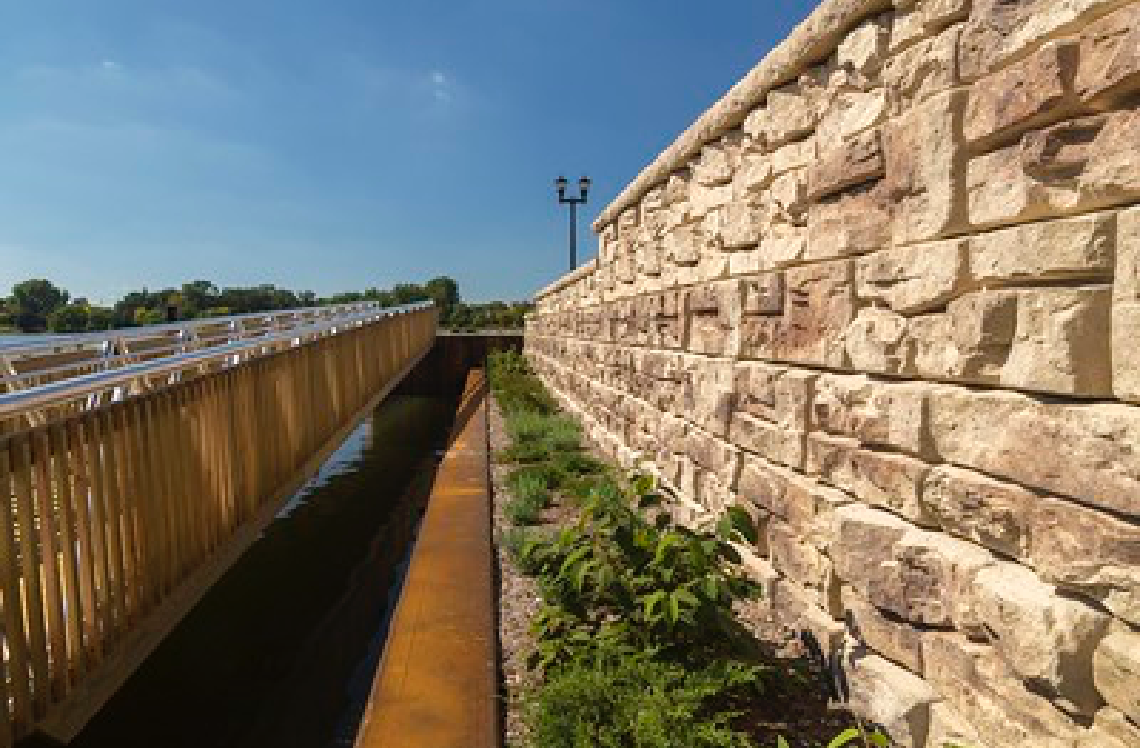 A stone wall along a river with a wooden bridge in the background.