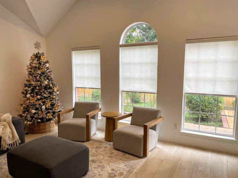 A living room with a Christmas tree has roller shades on the windows. 