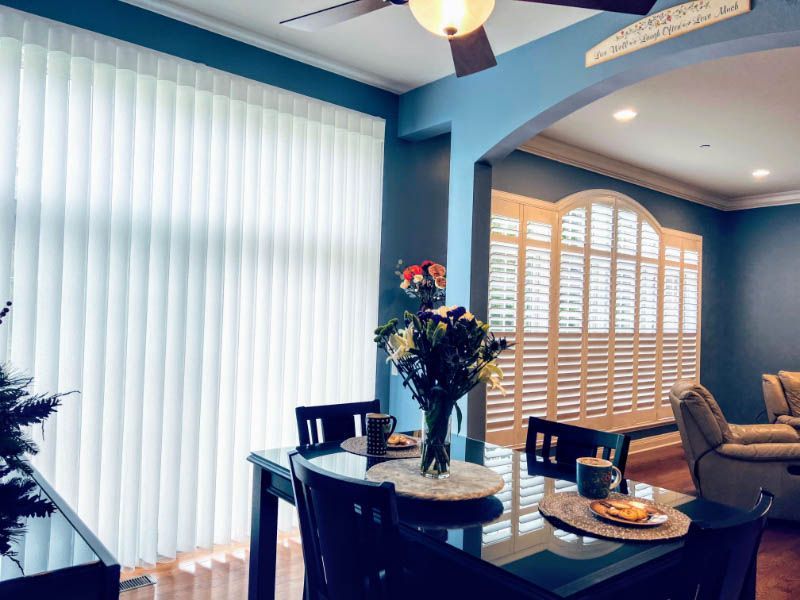 A blue dining room is shown with a window with shutters and glass doors with vertical shades.