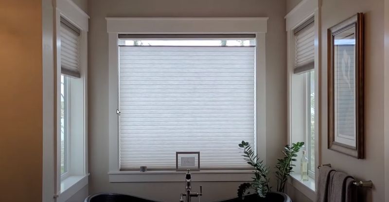 A bathroom has windows above the bath tub with cellular shades. 