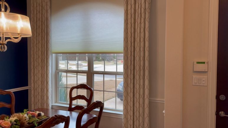 A dining room with a table and chairs and a window with drapery layered over roller shades. Love is Blinds