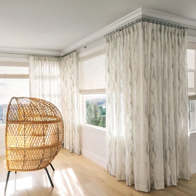 A living room with a wicker chair and white curtains layered over roller shades. Love is Blinds