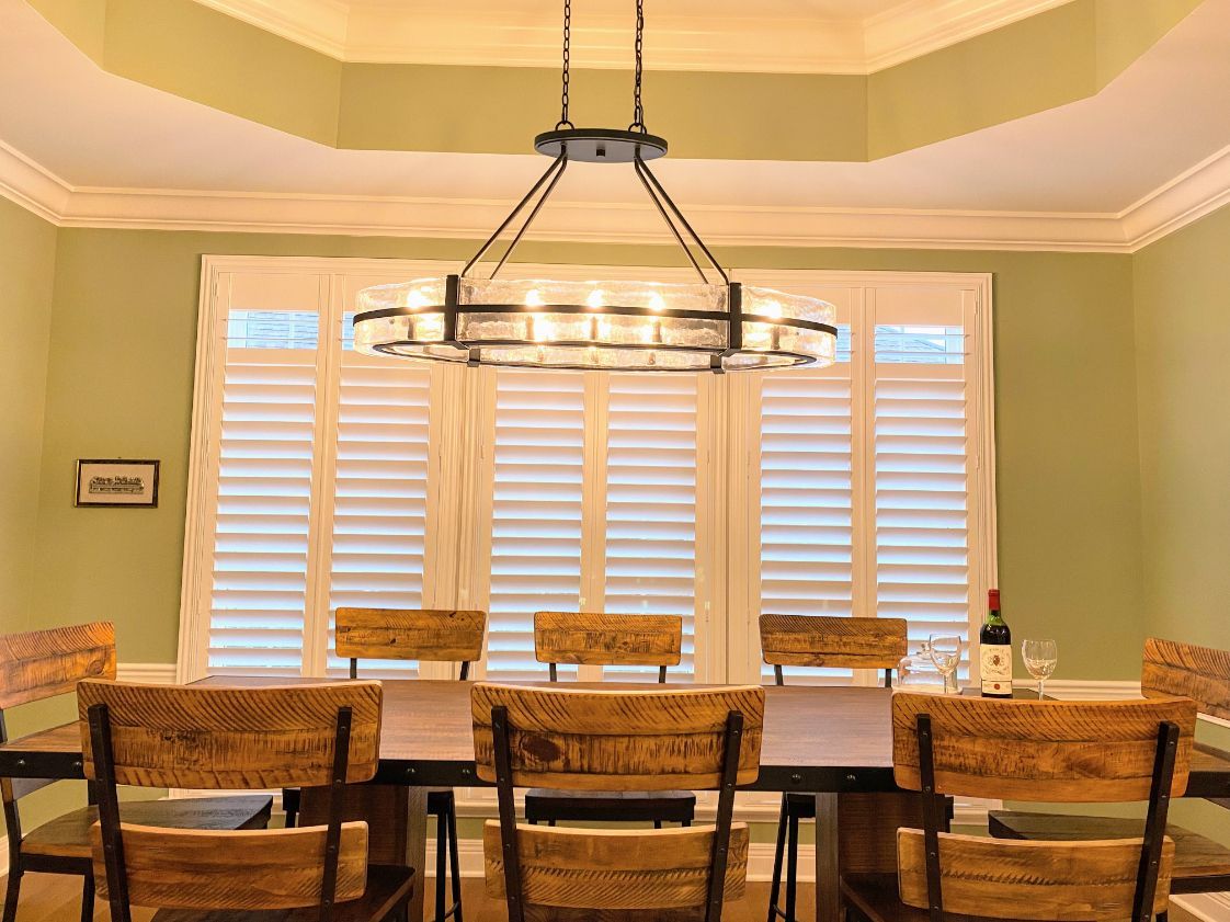 Dining room with wooden table, chairs, and large chandelier. Windows with shutters and light green walls.