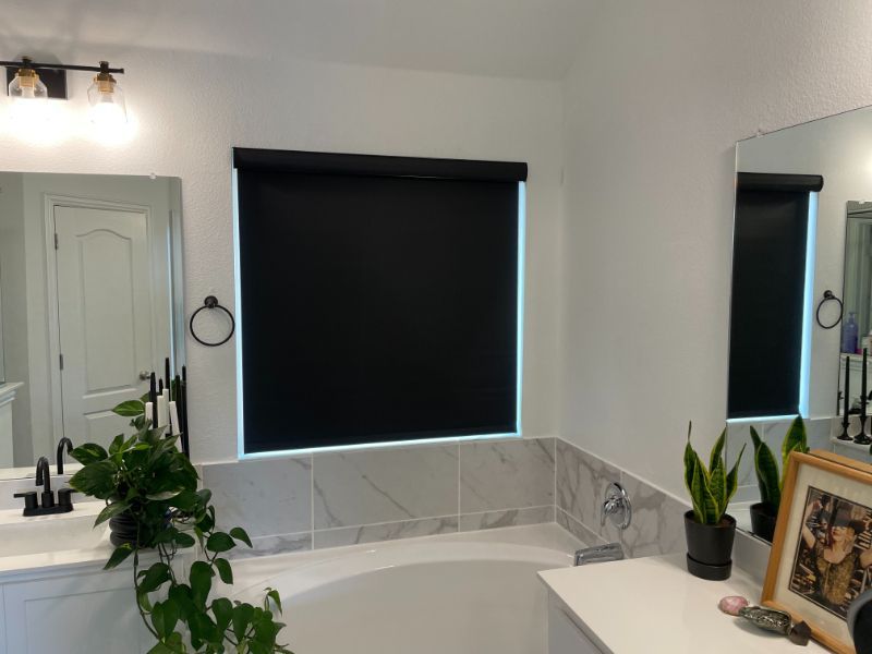 Love is Blinds TX: A bathroom with a tub , sink , mirror and black roller blinds.