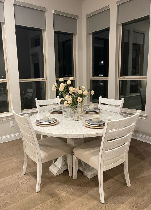 A dining room table and chairs with a vase of flowers on it. Roller shades on windows. 