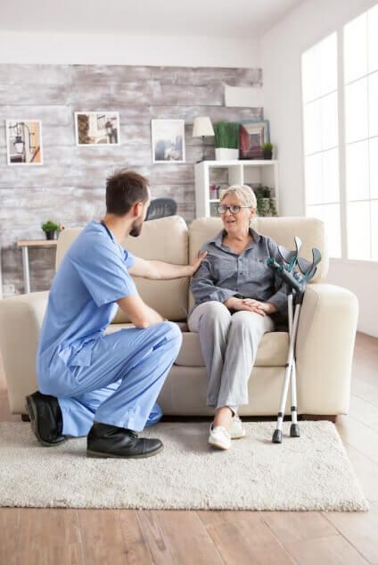 A nurse is kneeling down next to an elderly woman sitting on a couch.