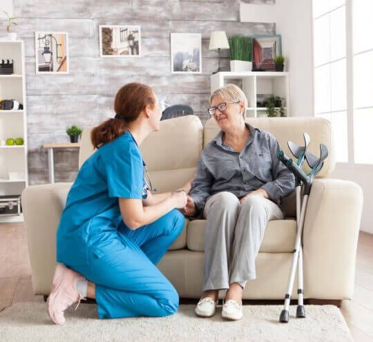 A nurse is kneeling down next to an elderly woman sitting on a couch.