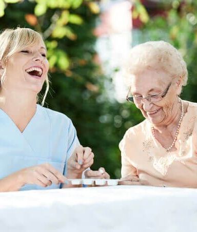 A nurse and an elderly woman are sitting at a table laughing.