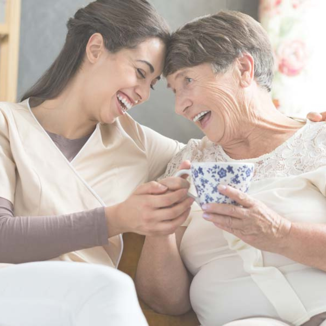 Two women are sitting next to each other holding cups of tea and smiling.